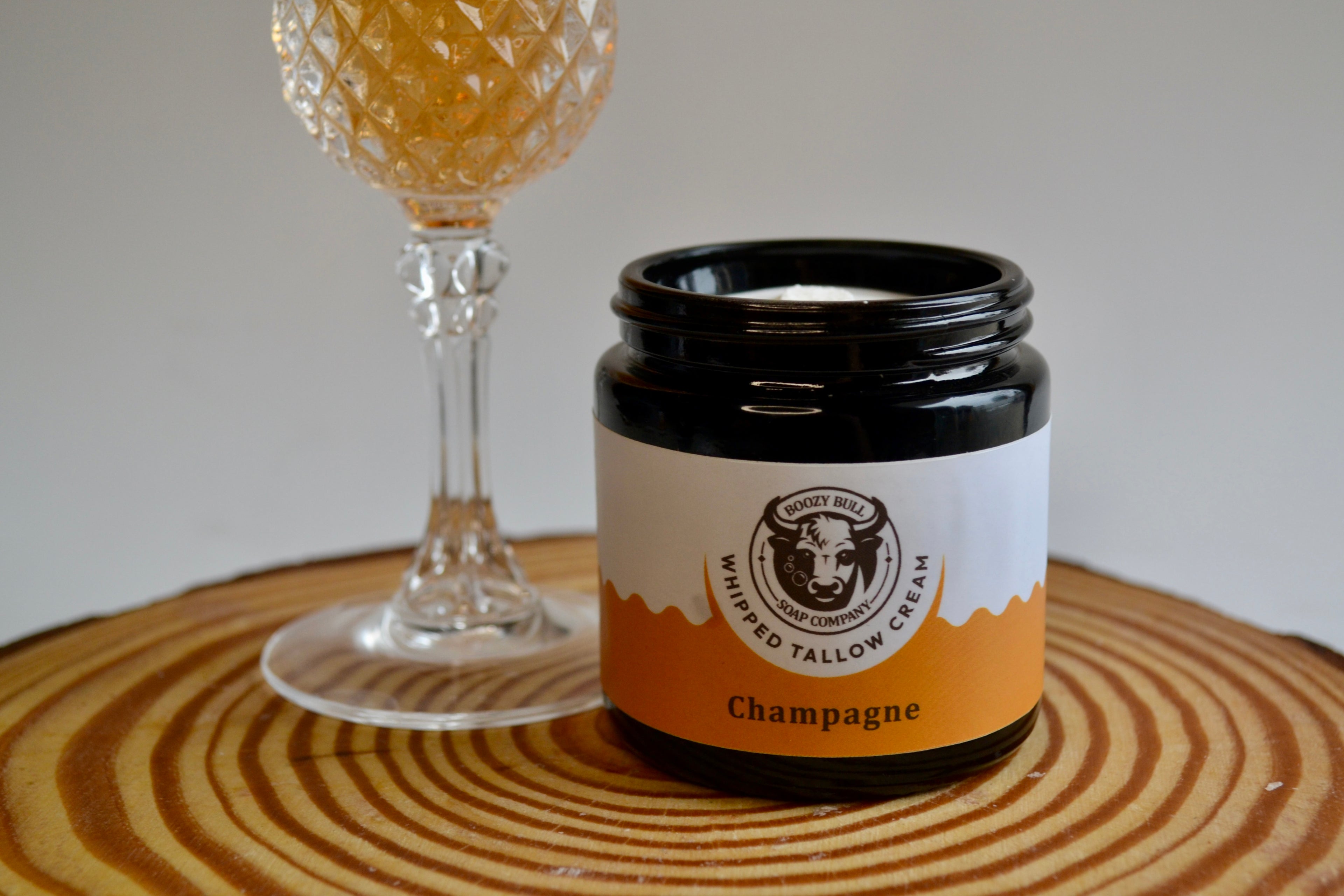 Jar of whipped tallow cream labeled 'Champagne' next to a glass on a wooden surface.