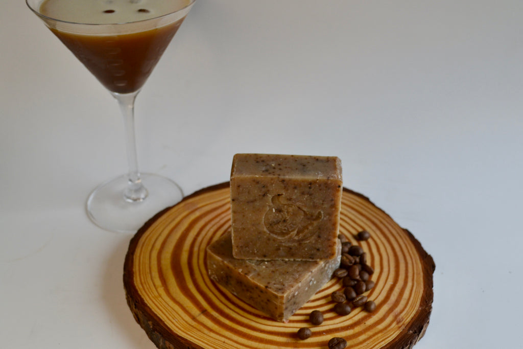 Two pieces of coffee-flavored soap on a wooden coaster with a glass of coffee in the background.