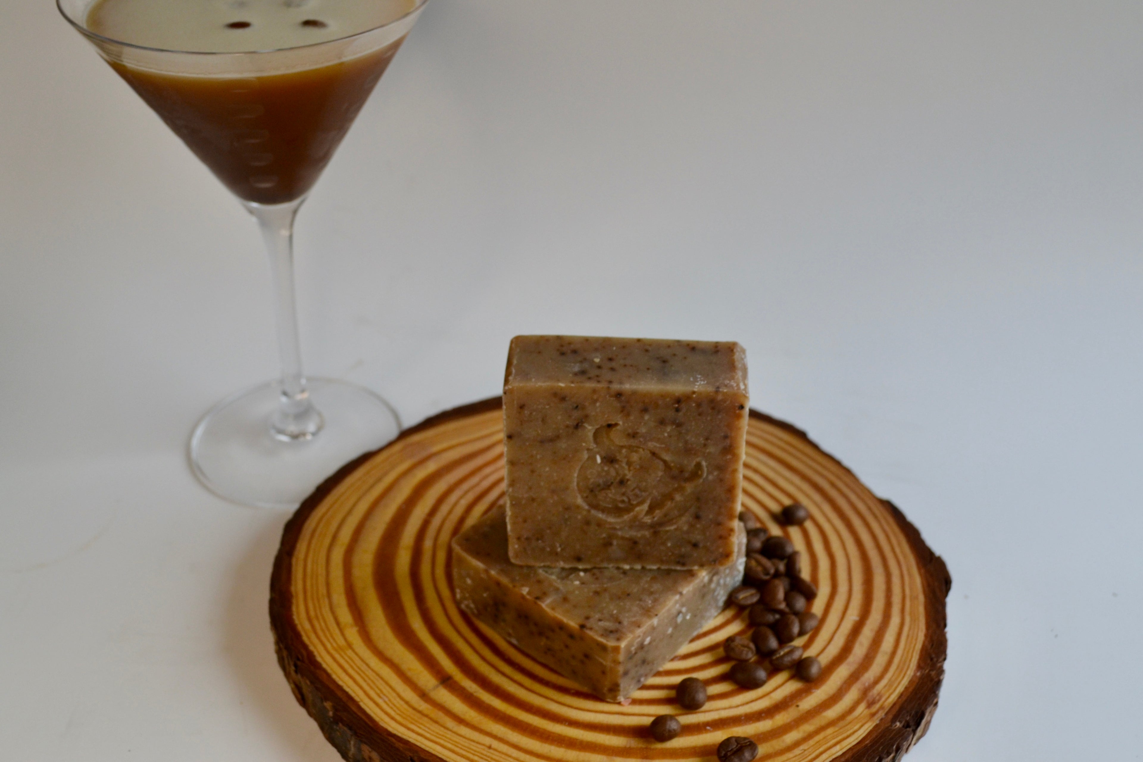 Two pieces of coffee-flavored soap on a wooden coaster with a glass of coffee in the background.