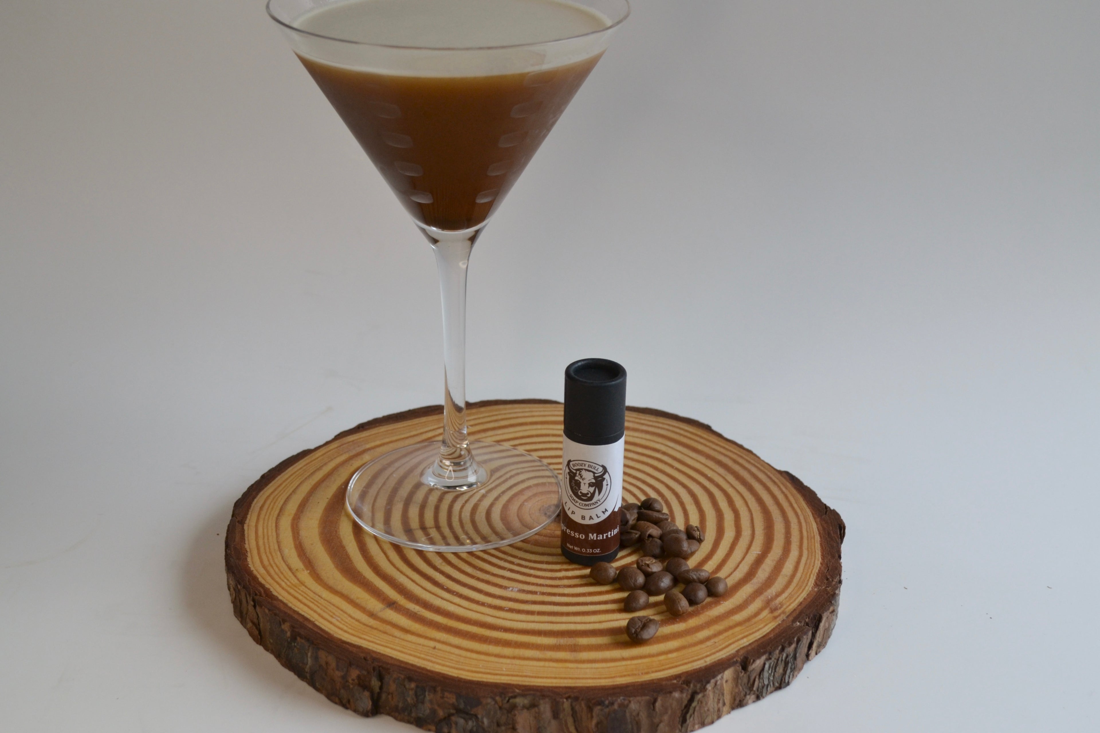 Cocktail glass with a dark liquid on a wooden coaster, accompanied by a small bottle and coffee beans.