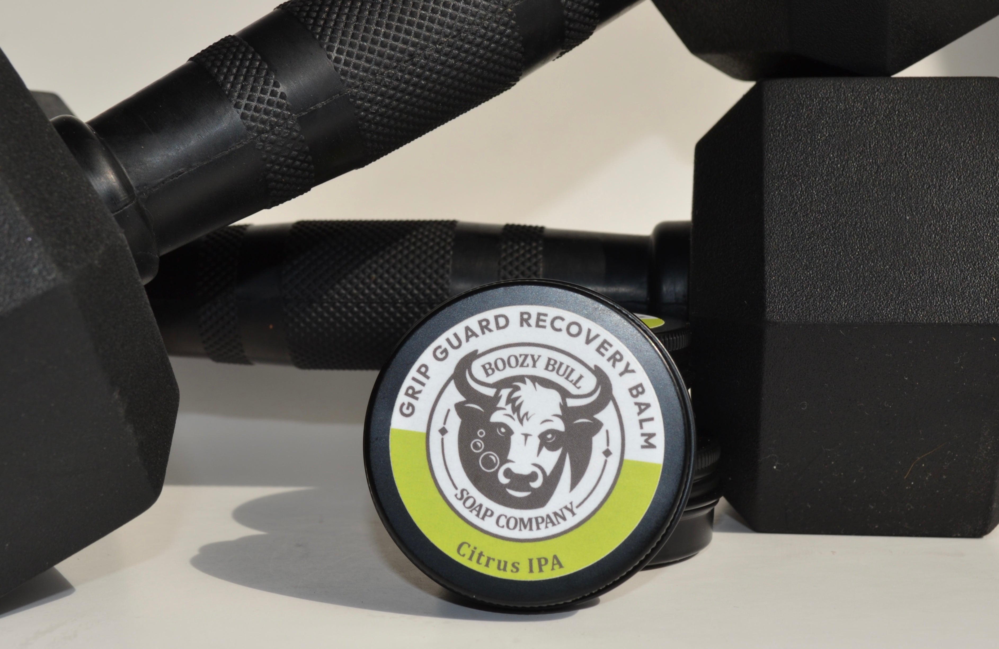 Grip recovery balm container with a cow logo on a surface with dumbbells.