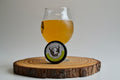 Glass of beer with a wooden coaster and bottle cap on a light gray background