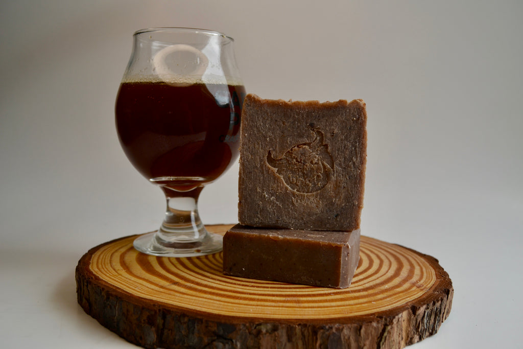 Two pieces of dark brown soap on a wooden block with a glass of dark liquid in the background.
