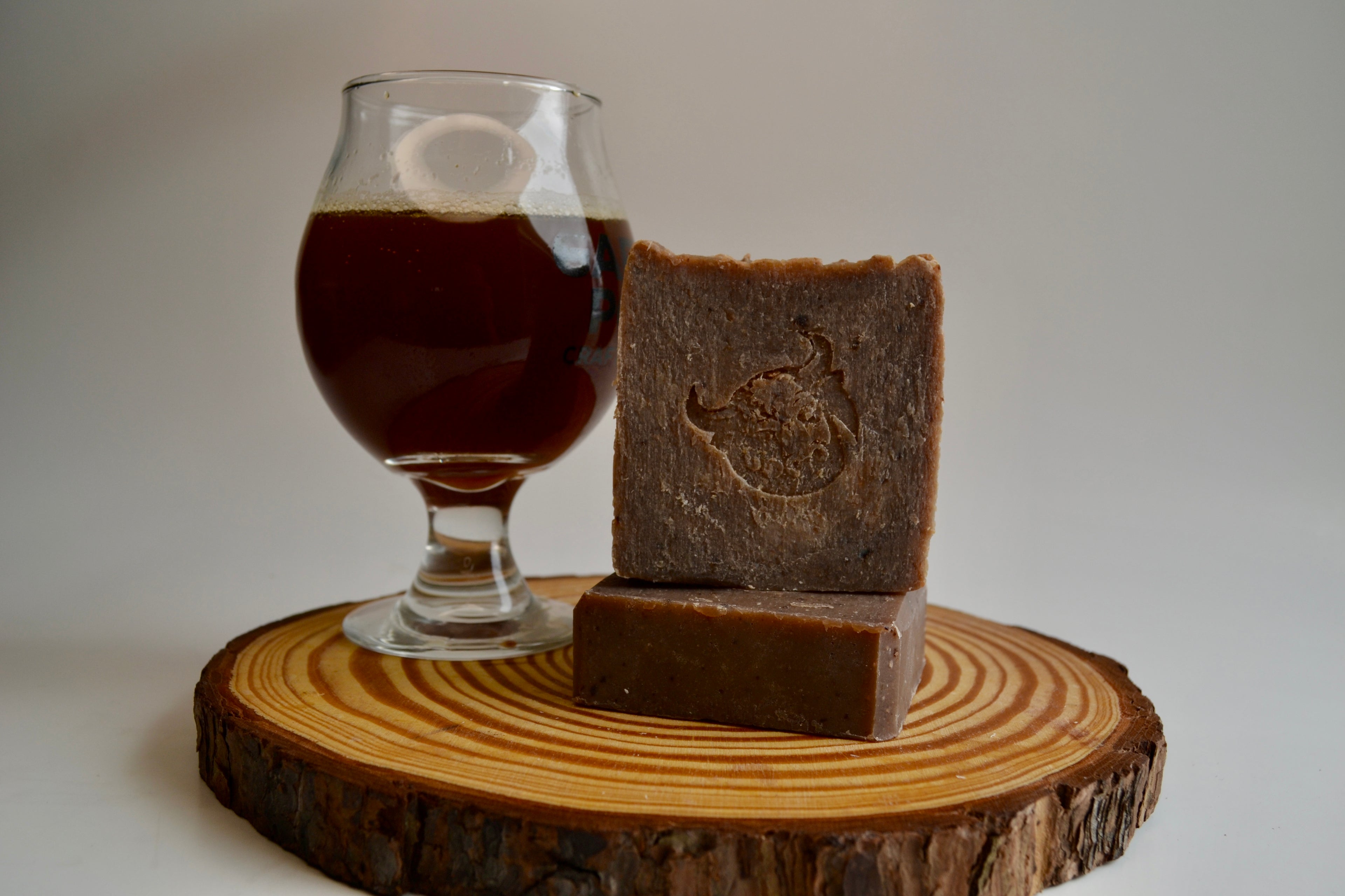 Two pieces of dark brown soap on a wooden block with a glass of dark liquid in the background.