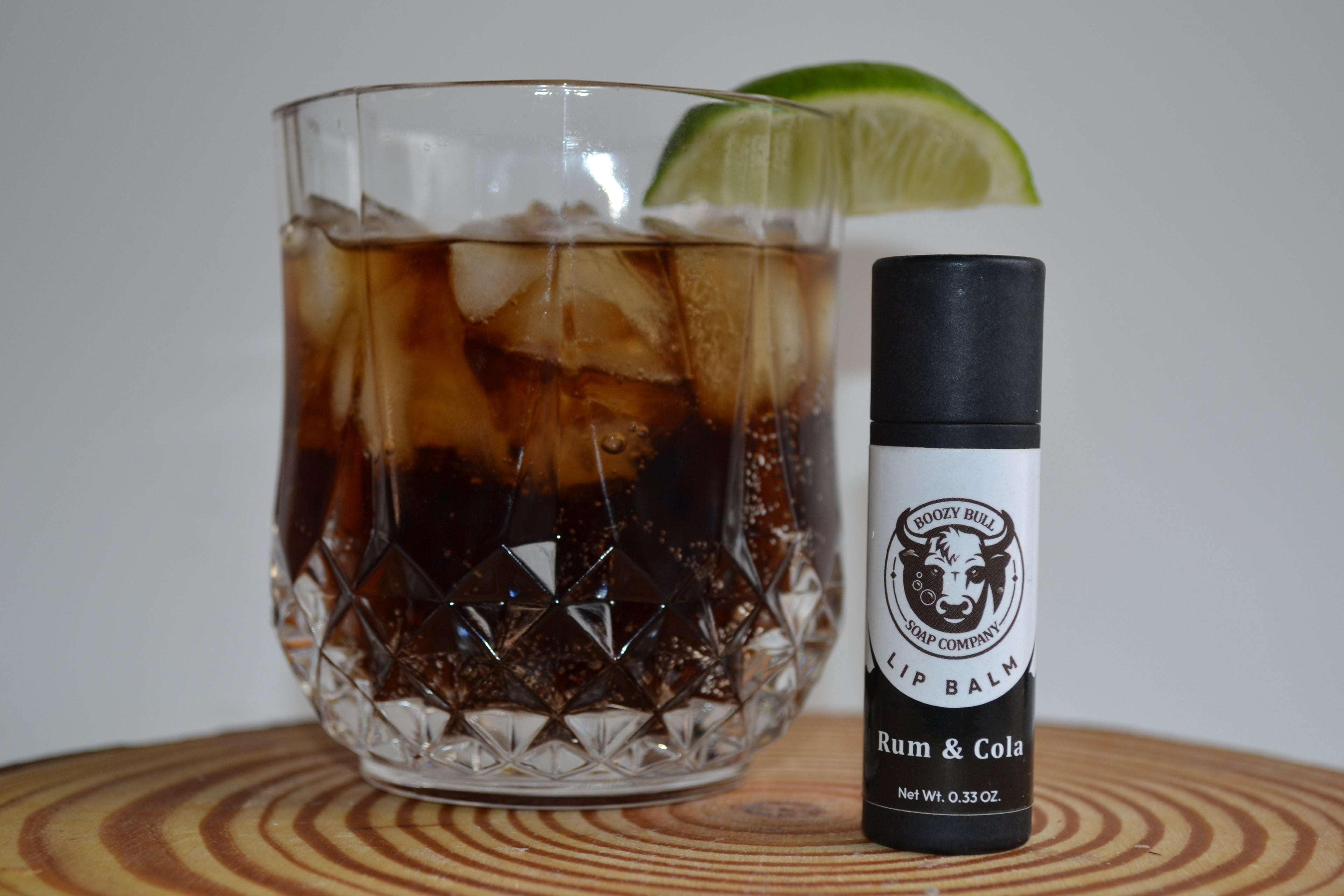 Glass of iced drink with a lime wedge and a bottle of 'Rum & Cola' lip balm on a wooden surface.