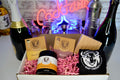 Gift set with bottles, a soap bar, and pink confetti in a box with a neon 'Cocktails' sign in the background.